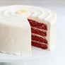 Daisy's Red Velvet Cake - A frosted red velvet cake on a white plate, with one slice cut out to show the layered red cake inside and a small yellow flower decoration on top.