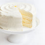 Daisy's Lemon Cake - A white frosted cake with a slice cut out, showing pale yellow layers, displayed on a white pedestal stand against a white background.