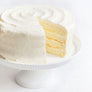 Daisy's Lemon Cake - A white frosted cake with a slice cut out, showing pale yellow layers, displayed on a white pedestal stand against a white background.
