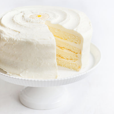 Daisy's Lemon Cake - A white frosted cake with a slice cut out, showing pale yellow layers, displayed on a white pedestal stand against a white background.
