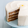 Daisy's Carrot Cake - A close-up of a layered carrot cake with white frosting, with a slice removed to show the interior. The cake is decorated with a small white daisy topper and sits on a light blue cake stand against a pale blue background.