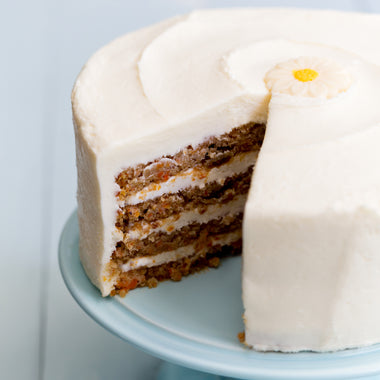 Daisy's Carrot Cake - A close-up of a layered carrot cake with white frosting, with a slice removed to show the interior. The cake is decorated with a small white daisy topper and sits on a light blue cake stand against a pale blue background.