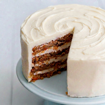 Daisy's Carrot Cake - A close-up of a layered carrot cake with white frosting, with a slice removed to show the interior. The cake sits on a light blue cake stand against a pale blue background.