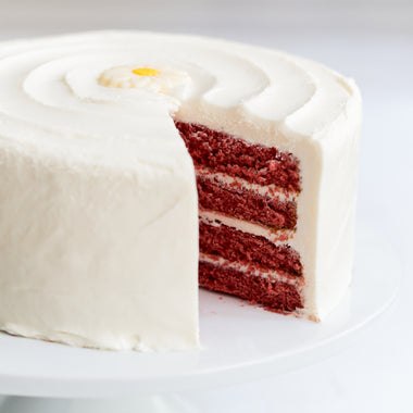 Daisy's Red Velvet Cake - A frosted red velvet cake on a white plate, with one slice cut out to show the layered red cake inside and a small yellow flower decoration on top.